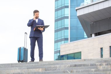 Kendine güvenen bir iş adamı, bir ofis binasının arka planında merdivenlerde duran bir bavulla duruyor.
