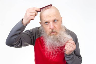 Kalın beyaz sakallı yaşlı kel adam, kırmızı tarak kullanıyor, diğer eliyle sakal çekiyor, kırmızı ve gri atletik gömlek giyiyor, beyaz arka plan.