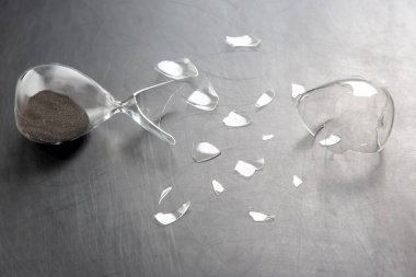 Parçalanmış cam parçalarıyla çevrili metalik bir yüzeye dökülen koyu kumlu kırık bir kum saati. Zaman, kırılganlık ve kayıp konuları için ideal..