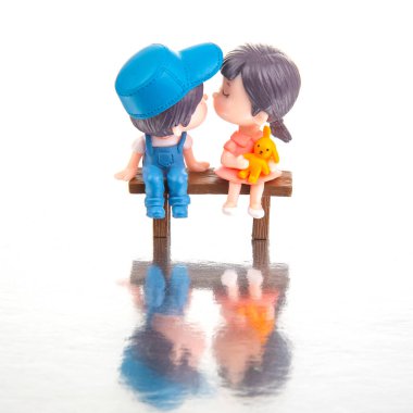 İki mini figür, mavi tulumlu bir çocuk ve pembe elbiseli bir kız, ahşap bir bankta yakın oturuyorlar. Durgun sulardaki yansımaları masumiyetin rüya gibi atmosferini yaratır..