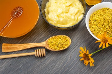 Bir kasede güzel bir bal aranjmanı, cam kasede kremsi bir madde, ve tahtadan bir masada ahşap bir kaşıkta arı poleni, canlı bir dokunuş için sarı çiçeklerle vurgulanmış..