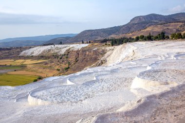 Pamukkale, mineral zengini termal sulardan oluşan eşsiz travertin teraslara sahiptir. Beyaz kalsiyum oluşumları, açık bir gökyüzünün altındaki yeşil manzaralar ve uzak dağlarla tezat oluşturuyor..