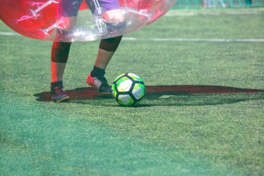 Katılımcılar parlak güneş ışığı altında iyi korunan bir çimenlikte balon futbol oynuyorlar. Bir oyuncu canlı bir futbol topunu açık şişme balon kıyafeti giyerken tekmelemeye hazır..