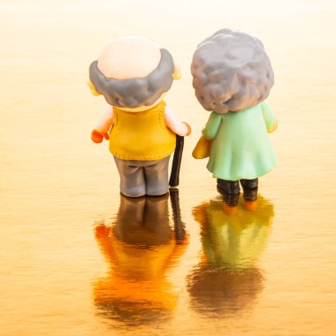 İki minyatür figür yan yana duruyor, parıldayan altın bir yüzeye bakıyor. Bir baston tutan yaşlı çift, arkadaşlığı ve deneyimleri paylaşmayı temsil ediyor..