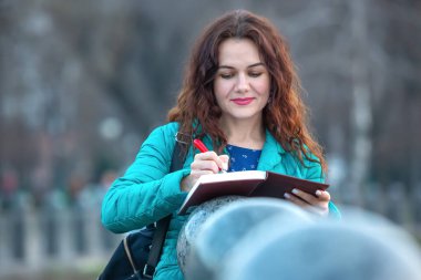 Uzun kıvırcık saçlı bir kadın deftere yazarken gülümsüyor. Şehir parkında korkulukların yanında duruyor. Yumuşak öğleden sonra ışığı odaklanmış ifadesini ve sakin atmosferini güçlendiriyor..