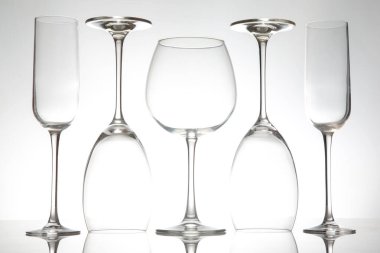 Flüt ve yuvarlak bir cam da dahil olmak üzere çeşitli cam şekillerden oluşan bir koleksiyon yumuşak beyaz bir arka plana karşı duruyor, parlak tasarımlarını ve yansıtıcı yüzeylerini sergiliyorlar..