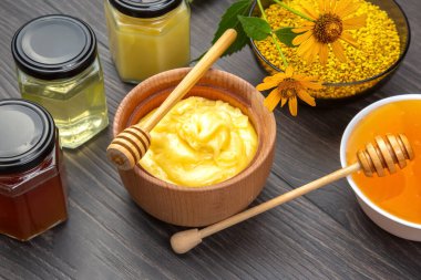 Doğal bal, balmumu ve canlı çiçekler, bal kavanozlarıyla dolu ahşap bir masada güzelce düzenlenir. Bal aygırı bir kase bal içinde dinleniyor, tatlı, altın dokular sergiliyor.