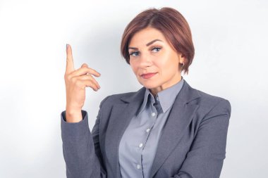 Kendine güvenen bir iş adamı, gri bir takım elbise ve gömlek giymiş, iyi ışıklandırılmış bir stüdyoda duruyor, tek parmakla işaret ediyor, vurgu yapıyor veya güçlü bir fikir sunuyor..