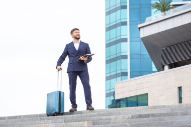Resmi giyinmiş bir adam, çağdaş bir binanın dışındaki merdivenlerde güven içinde duruyor. Bir elinde bir tablet, diğer elinde bir bavul var. Hareketli şehirde yolculuğuna hazır..