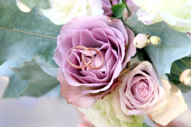 Lavanta ve krem güllerle süslenmiş güzel bir düğün buketi. Altın alyanslar çiçeklerin üzerinde, sevgi ve bağlılığı sembolize ediyor. Ayarlama samimi bir anı yakalar..