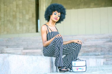 Afrikalı bir kadın, kültür merkezinde taş basamaklarda otururken şık çizgili tulumuyla moda anlayışını sergiliyor. Cesur makyajı ve eşsiz saç şekli kendine güvenini arttırıyor..