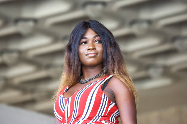 A woman of African descent stands proudly, showcasing her fashion sense in a vibrant attire. The background features a modern architectural design, highlighting a sunny day ambiance.