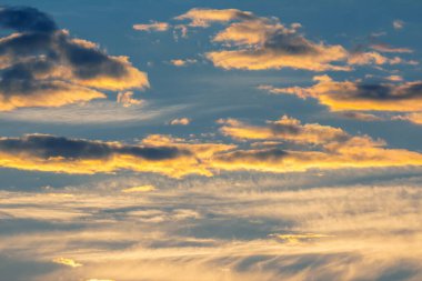 Altın ve turuncu renkler, gün batımında gökyüzünde sürüklenen bulutlar gibi mavi tonlarla harmanlanır. Çarpıcı renkler, alacakaranlıkta sakin ve huzurlu bir atmosfer yaratıyor. Bu da günün sonunu işaret ediyor..