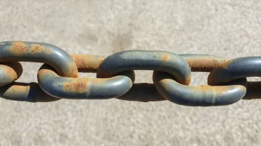 Paslı metal bir zincirin yakın çekim görüntüsü, aşınmış halkaların detaylarını ve farklı renkleri gösteriyor, altında desenli bir yüzey, zamana ve elementlere maruz kalma.