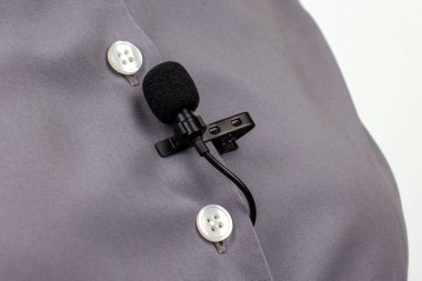 Gri bir gömleğe takılı yakalı bir mikrofonun yakın plan görüntüsü, röportaj, sunum ya da videolar sırasında ses yakalama tasarımını gösteriyor.