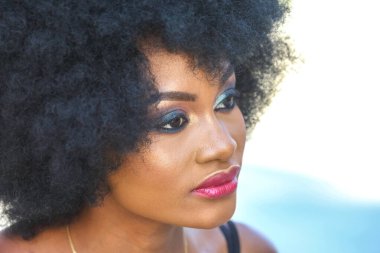 Genç bir Afrikalı kadın açık havada güneşli bir günün tadını çıkarırken canlı doğal saçlarını ve ustalıkla sürdüğü makyajını sergiliyor. Kendine güveni ve ışıl ışıl cildi güzelliğini vurguluyor..