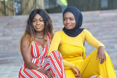 İki Afrikalı kadın açık havada canlı bir yerde oturuyorlar, parlak ve şık kıyafetler giyiyorlar. Bir kadın çizgili bir elbise giyerken diğeri sarı bir tulum ve başörtüsü takıyor..