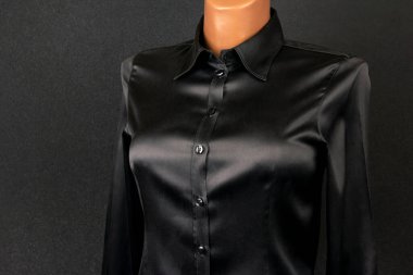 Pürüzsüz, siyah, saten, düğmeli bir gömlek, koyu renk bir arka plana karşı bir mankenin üzerinde sergileniyor, parlak ve şık tasarımını ön plana çıkarıyor. Resmi etkinlikler ya da şık günlük kıyafetler için ideal kıyafet.