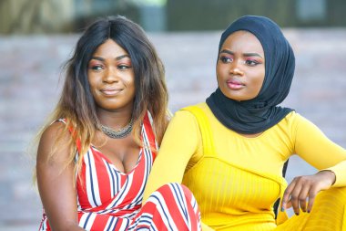 İki Afrikalı kadın yan yana oturuyor, biri çizgili elbiseli, diğeri sarı elbiseli, siyah başörtülü, gülücükler ve açık hava ortamında sohbet ediyorlar..