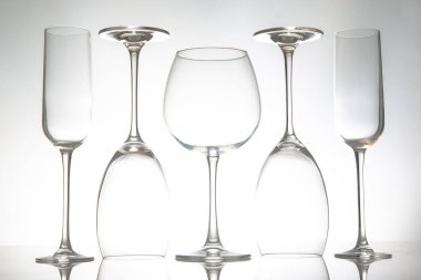 Uzun saplı beş zarif, boş şarap kadehinden oluşan gösterişli bir sıra yumuşak gri arka planda parlak bir yüzeye yansıyor. Yemek ya da etkinlik temaları için mükemmel..