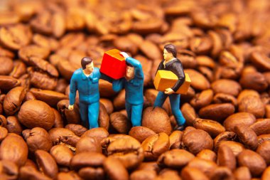 Mavi üniformalı üç minyatür figürden oluşan yaratıcı bir yakın plan kahve çekirdekleri denizinin ortasında ticaret ve ticareti sembolize eden sarı bir kutu ve kırmızı paket değiş tokuşu. Sıcak kahverengi tonlar zengin, dokulu bir zemin oluşturur..