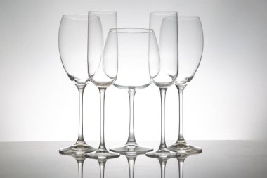 İnce saplı beş boş şarap kadehinin minimalist görüntüsü, yansımaları yumuşak gri bir arkaplana karşı parlak bir masada simetri yaratıyor..