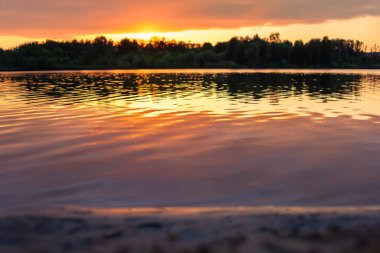 Huzurlu bir göl üzerinde sersemletici bir gün batımı, dalgalı sulara canlı turuncu renkler yansıtıyor, bir orman silueti tarafından çerçevelenmiş. Doğa ve manzara fotoğrafçılığı için ideal..