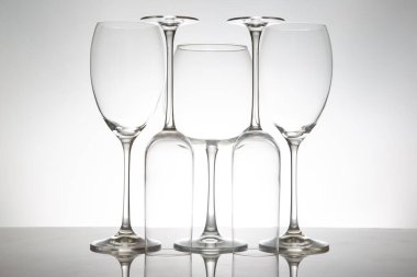 İnce saplı beş boş şarap kadehinin minimalist görüntüsü, yansımaları yumuşak gri bir arkaplana karşı parlak bir masada simetri yaratıyor..