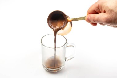 Bakır bir bardaktan cam bir fincana dökülen Türk kahvesinin yakın çekimi, kremalı köpüğünü ve beyaz arka planda koyu rengini gözler önüne seriyor.