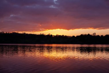 Gün batımında dingin göl, sakin sularda altın yansımalar, çarpıcı bir gökyüzünün altında yemyeşil ağaçlarla çevrili. Doğa ve sükunet temaları için mükemmel.