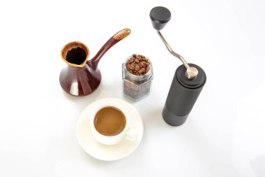 Siyah manuel kahve öğütücüsü, ahşap saplı seramik bir taş ve beyaz arka planda demlenmiş beyaz bir fincan kahve etrafını saran kavrulmuş kahve çekirdekleri..