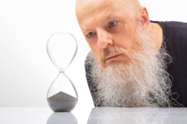 Beyaz sakallı kel bir adam zamanın sonsuz akışını simgeleyen kum saatini yakından inceliyor. Fotoğrafın minimalist tarzı, içgözlem duygusunu ve zamanın akışını arttırır.
