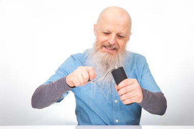 Uzun beyaz sakallı neşeli kel bir adam, el değirmeni ile kahve çekirdeklerini öğütür, gri kollu mavi bir kot gömlek giyer, düz beyaz bir arka plan üzerine kuruludur