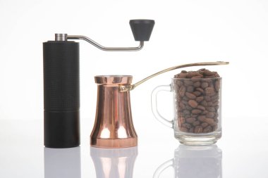 Sade bir kahve öğütücüsünün yakın çekimi, parlak bir bakır çakmak, ve kahve çekirdekli cam bardak, kahve hazırlama görselleri için temiz beyaz bir zemin üzerine kurulmuş.