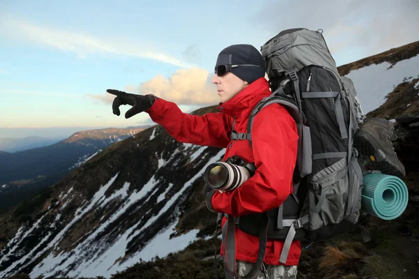 Ο ταξιδιώτης στο κόκκινο σακάκι με σακίδιο και φωτογραφική μηχανή στο χέρι σημεία σε την απόσταση χέρι είναι στην πλαγιά του λόφου — Φωτογραφία Αρχείου