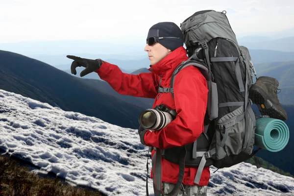 Путешественник в красной куртке с рюкзаком и камерой в руке указывает на дистанционную руку, находящуюся на склоне холма — стоковое фото