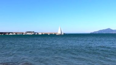 Beyaz yelkenli yelkenli, güneşli bir günde mavi denizde marinadan ayrılıyor. Doğa ve ulaşım. Dinlenme ve turizm. - Evet. Yüksek kalite 4k görüntü