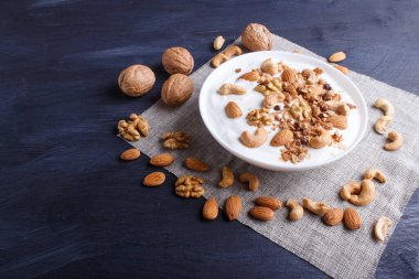 Yunan yoğurtlu beyaz tabak, granola, badem, ceviz, siyah ahşap arka planda ceviz. Kapat. Boşluğu kopyala.