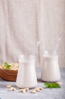 Sütsüz organik kaju sütü bardakta ve fındıklı ahşap tabakta gri beton arka planda. Vejetaryen sağlıklı gıda konsepti, yakın plan, yan görüş..