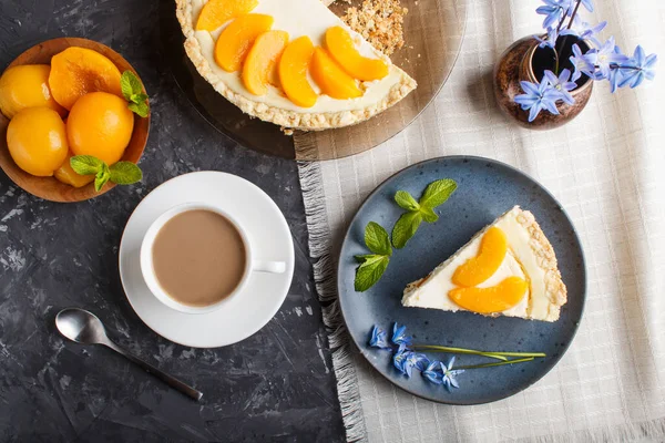 A piece of peach cheesecake on a blue ceramic plate with blue flowers and a cup of coffee on a linen napkin on a black concrete background. top view, close up.