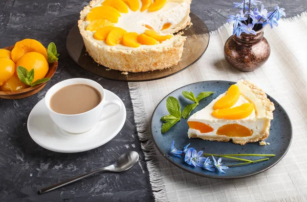 A piece of peach cheesecake on a blue ceramic plate with blue flowers and a cup of coffee on a linen napkin on a black concrete background. side view, close up.