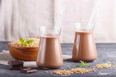 Süt ürünü olmayan organik yulaf ezmesi, bardakta çikolatalı süt ve siyah beton arka planda yulaf tohumlu ahşap tabak. Vejetaryen sağlıklı gıda konsepti, yakın plan, yan görüş..