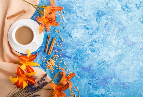 Orange day-lily and lavender flowers and a cup of coffee on a blue concrete background, with orange textile. Morninig, spring, fashion composition. Flat lay, top view, copy space.