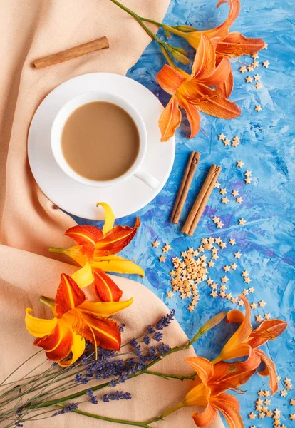 Orange day-lily and lavender flowers and a cup of coffee on a blue concrete background, with orange textile. Morninig, spring, fashion composition. Flat lay, top view, close up.