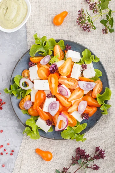 Vegetarian salad with fresh grape tomatoes, feta cheese, lettuce and onion on blue ceramic plate on gray concrete background and linen textile. top view, flat lay, close up.