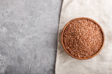 Ahşap kase, gri beton arka planda cilalanmamış kahverengi pirinç ve keten tekstil. Üst görünüm, düz görünüm, kopyalama alanı.