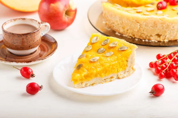 Traditional american sweet pumpkin pie decorated with hawthorn red berries and pumpkin seeds with cup of coffee on a white wooden background. side view, close up, selective focus.