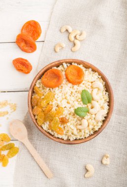 Beyaz ahşap bir arka plan ve keten tekstil üzerinde kurutulmuş kayısı, kuru üzüm ve kaju fıstığı ile bulgur lapası. Üst manzara, düz yatak, yakın çekim. Türk geleneksel mutfağı.