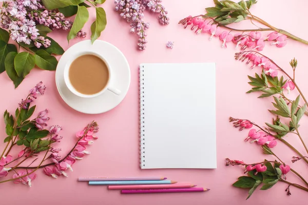 Mor leylak ve kanayan kalp çiçekleri ve bir fincan kahve ile defter ve pastel pembe arka planda renkli kalemler. Morninig, bahar, moda kompozisyonu. Düz yatış, üst görünüm.