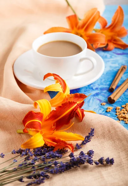 Orange day-lily and lavender flowers and a cup of coffee on a blue concrete background, with orange textile. Morninig, spring, fashion composition. side view, selective focus.
